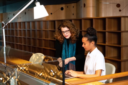 Woman gesturing to the screen of a laptop being used by a young person. A lamp is pointed towards them and behind them are wooden shelves.