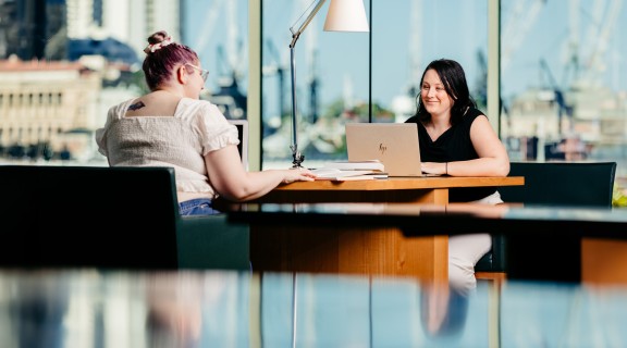 Two women sitting at a joint desk with a view of Brisbane city using laptops at State Library.